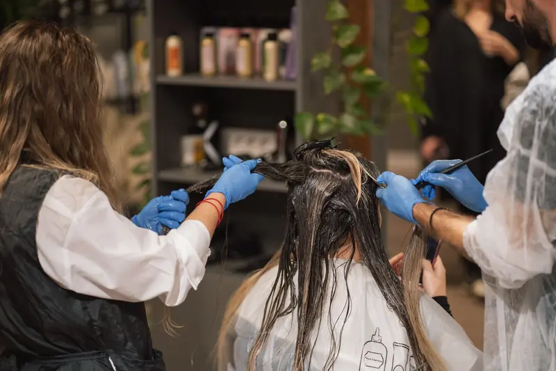 Dos estilistas con guantes aplicando color en el pelo de una clienta.