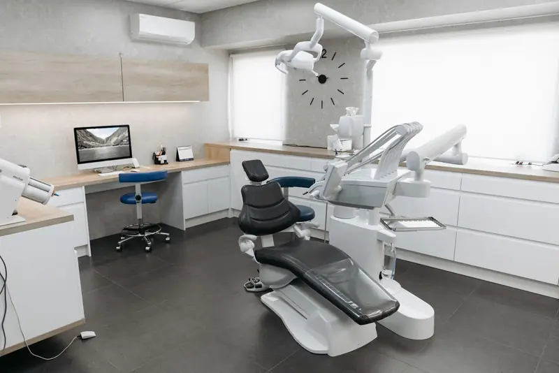 Consultorio con sillón odontológico gris, lámpara y mesada blanca.